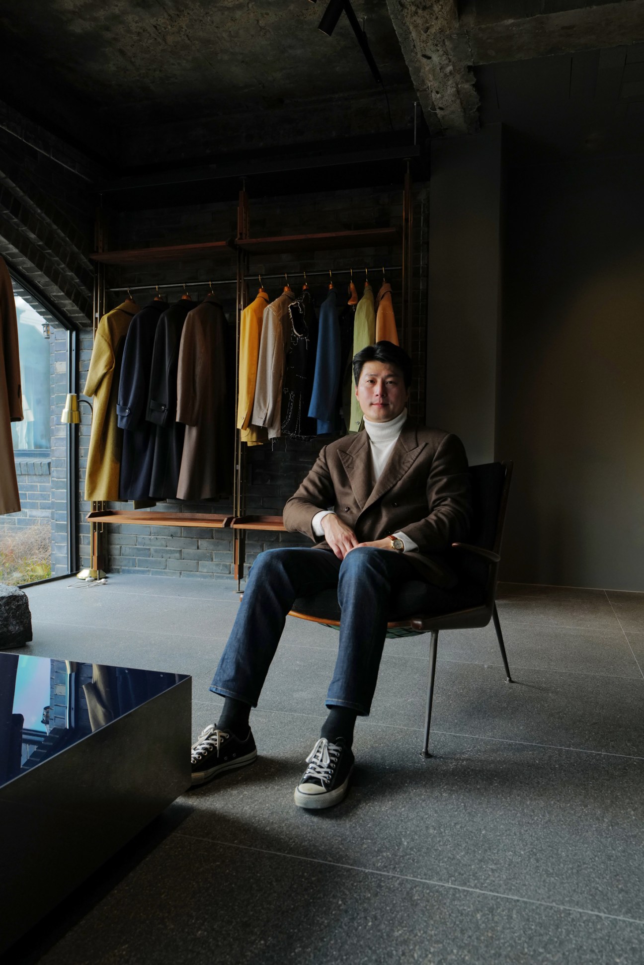 Kim Daechul sits in a black chair wearing a brown blazer in a minimalist room with clothing hanging on a rack behind him