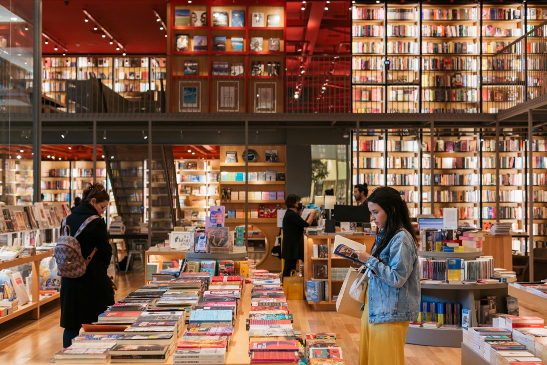 Modern bookstore interior with floor-to-ceiling bookshelves, customers browsing books on wooden display tables