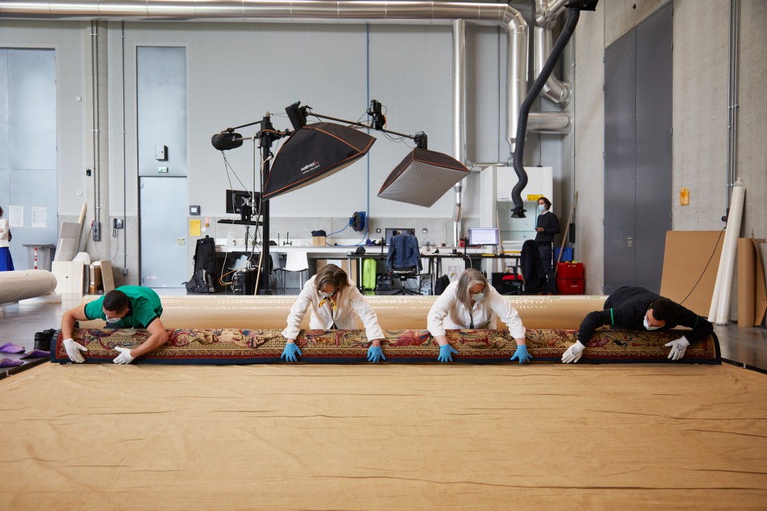 Restorers examine and preserve a large historical tapestry laid out on the floor in a bright conservation studio.