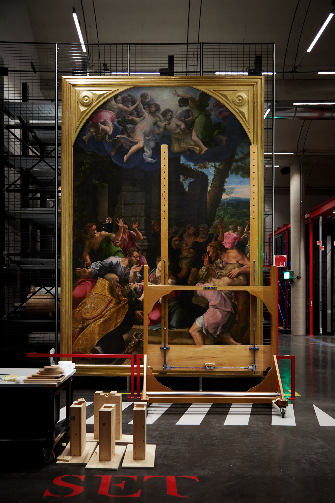 A large religious painting with gold frame mounted on protective wooden supports in a modern industrial storage facility.