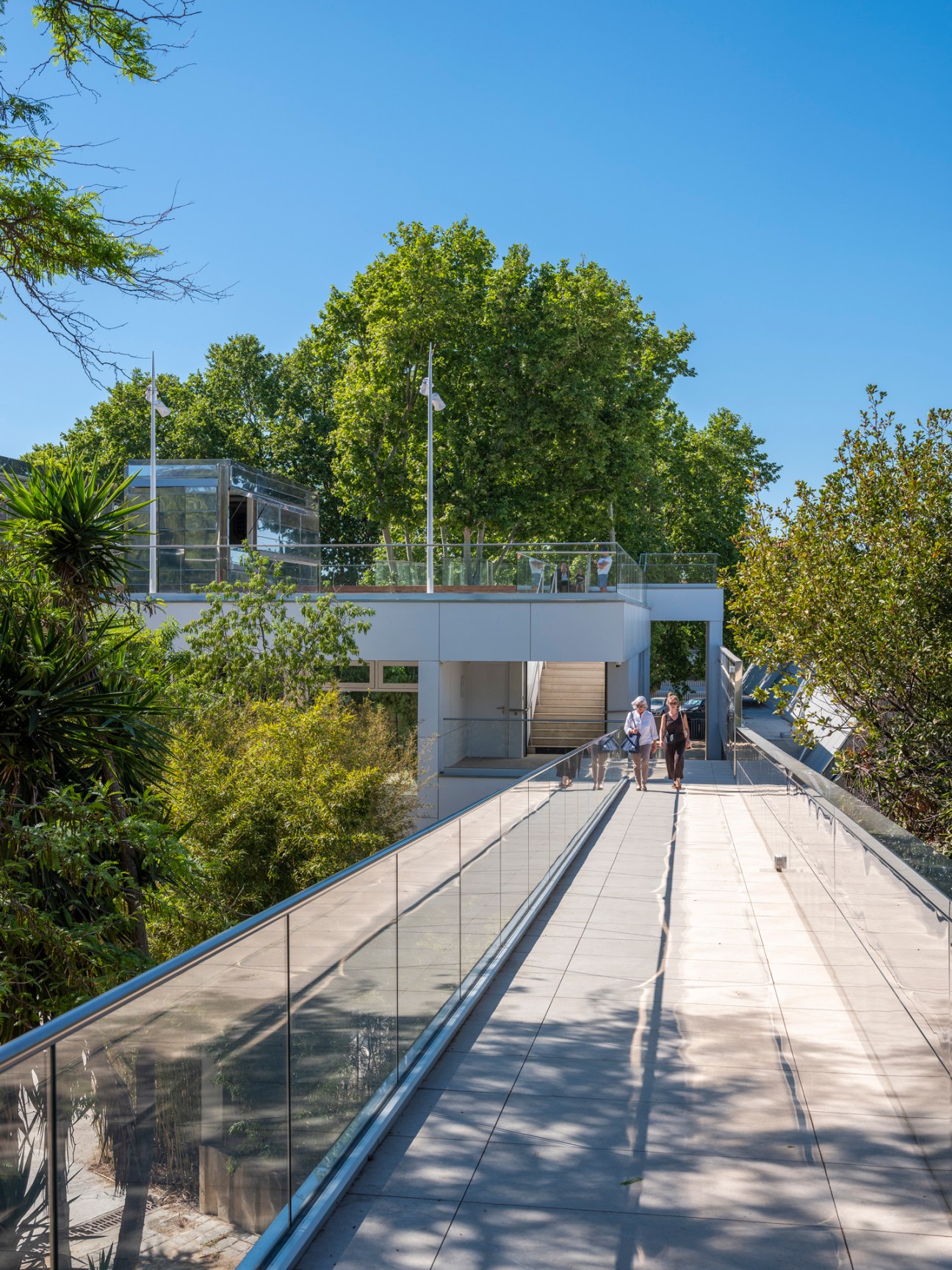 Modern white building with glass railings and walkway surrounded by Mediterranean vegetation.