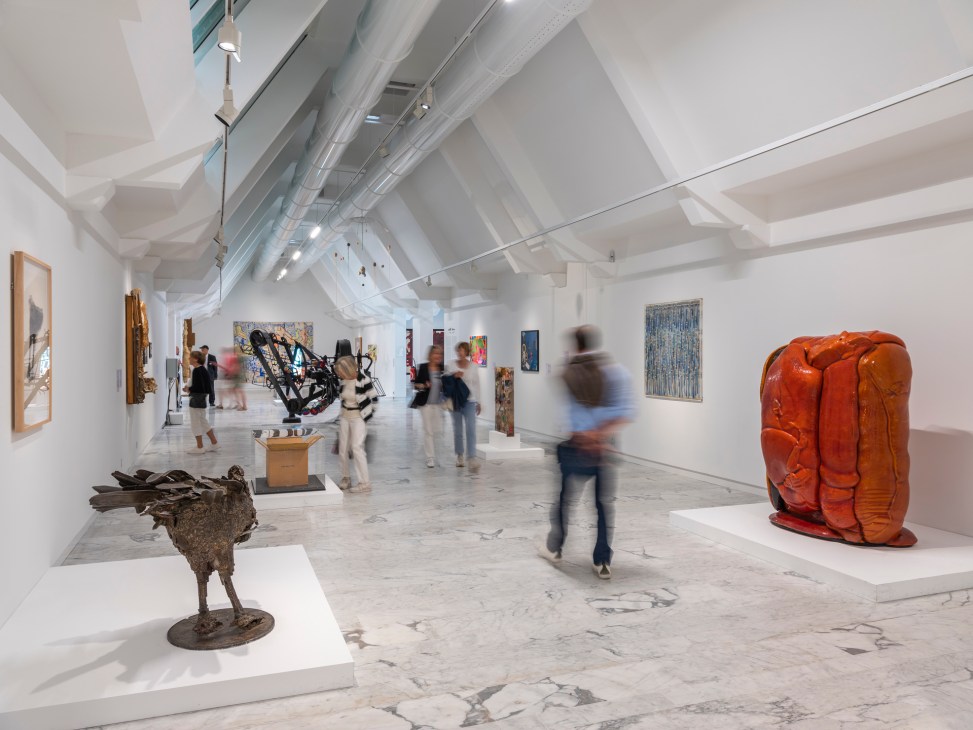 Gallery interior with white walls, skylights, sculptures on pedestals and paintings displaying contemporary art.