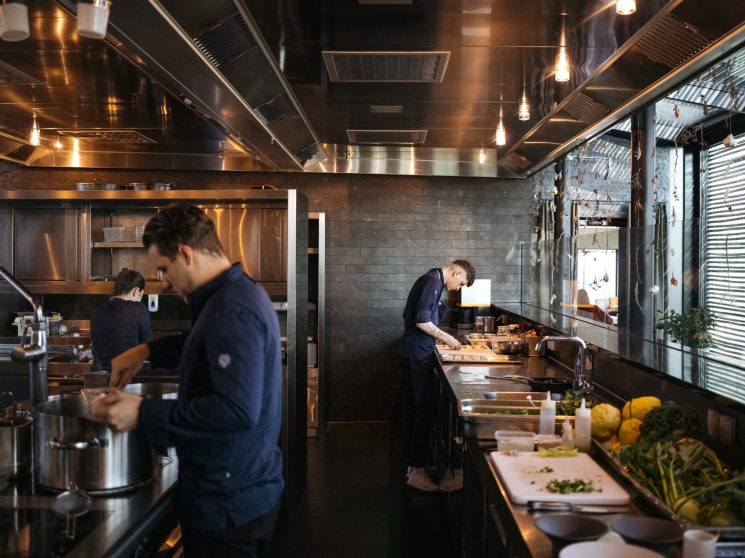 Modern restaurant kitchen with glass walls, stainless steel equipment, and chefs preparing food in professional setting