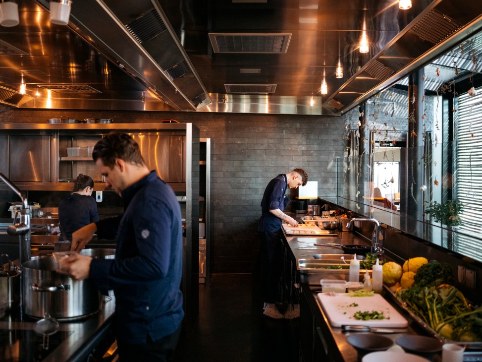 Modern restaurant kitchen with glass walls, stainless steel equipment, and chefs preparing food in professional setting