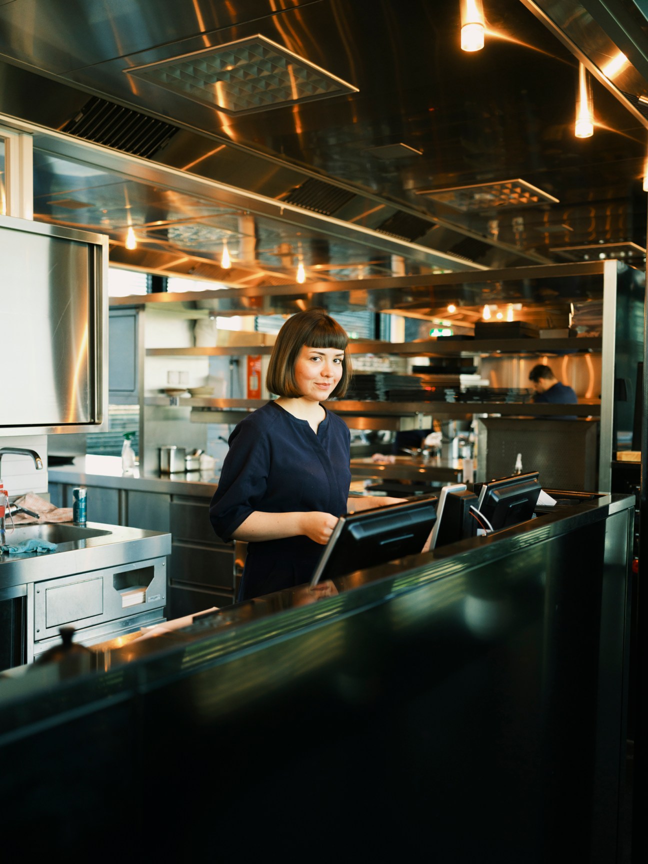 Woman with short brown hair in dark blue uniform working at point-of-sale system in modern restaurant kitchen