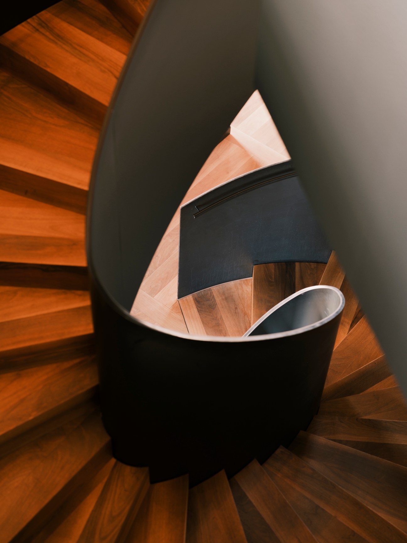 Curved spiral staircase with wooden steps and black bathtub leading downward
