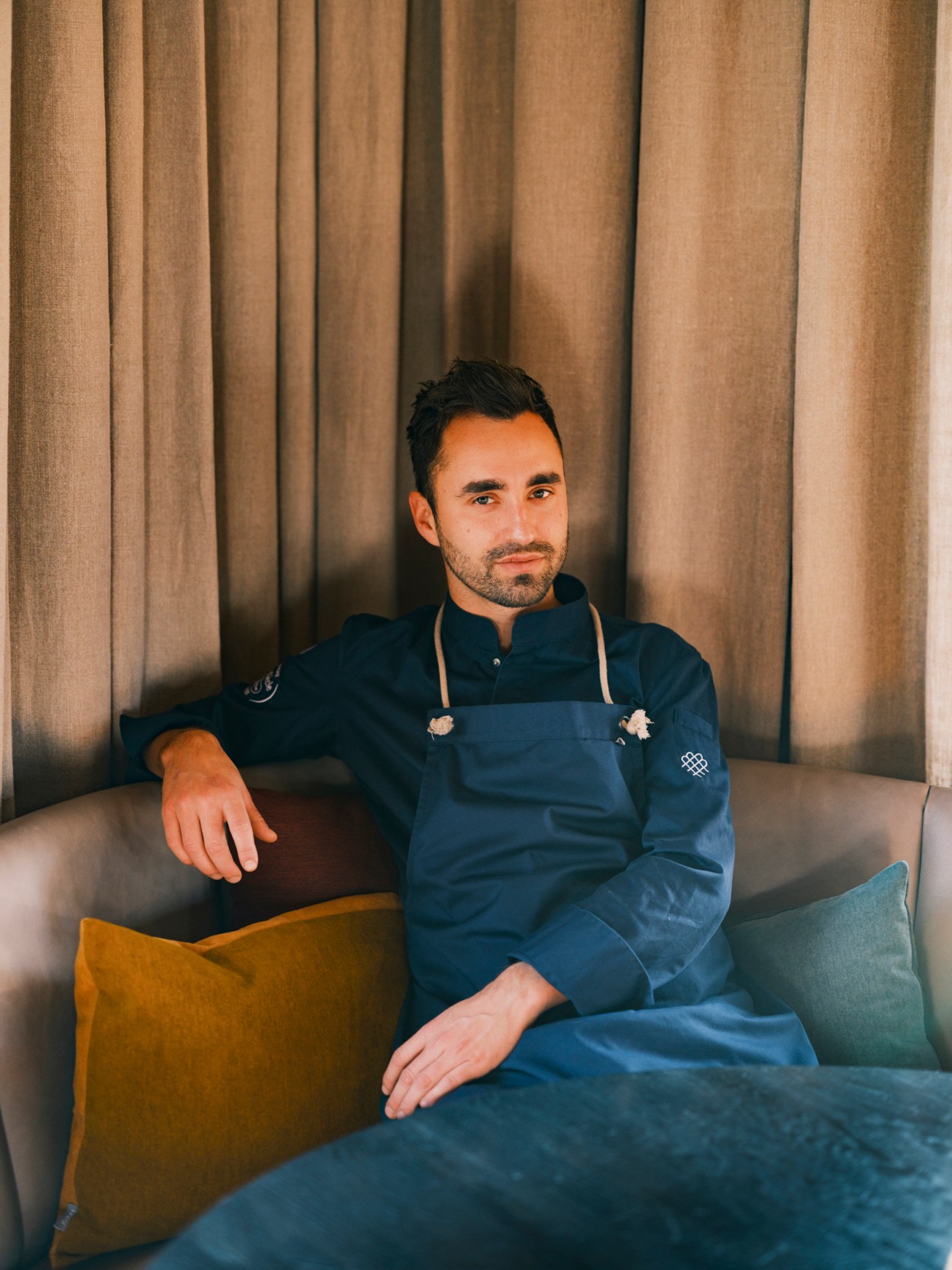 Chef Silvio Germann in blue chef's jacket sitting on grey sofa in restaurant lounge
