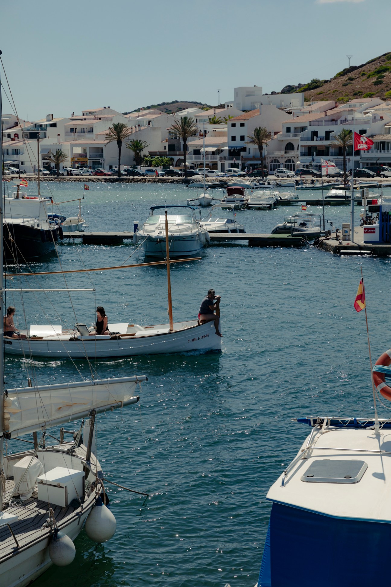 Moored sailing boats in a Mediterranean harbor with white waterfront buildings and palm trees on the hillside.