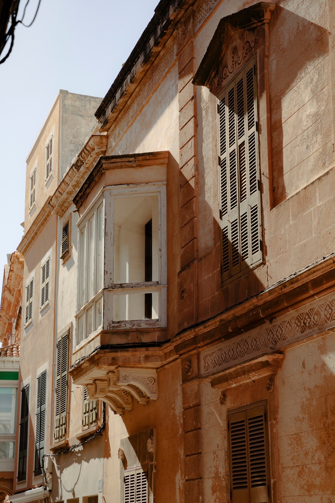 Weathered stone facade of a traditional Menorca building with white shuttered windows and ornate brown wooden details.
