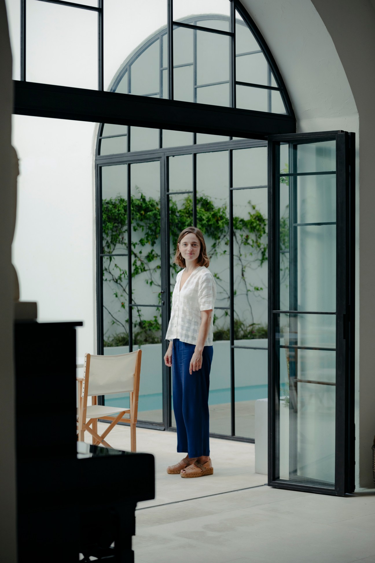 Woman in white shirt and navy trousers standing in modern greenhouse with black-framed glass doors and climbing ivy.