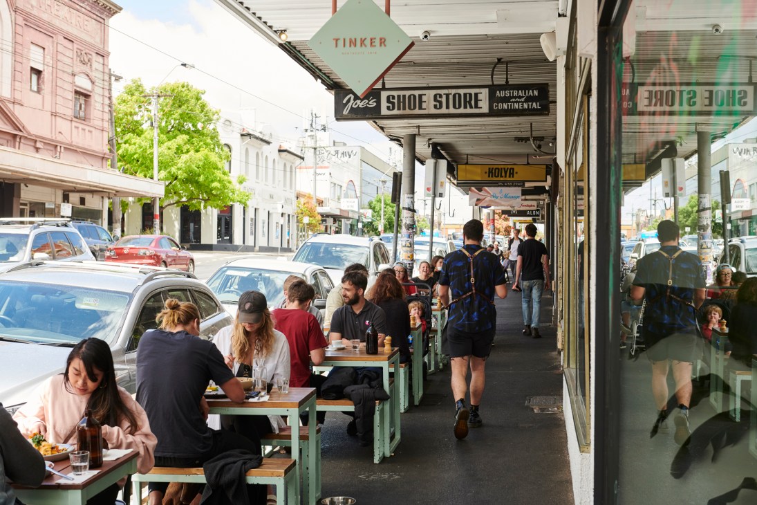 Diners seated at outdoor tables along a Melbourne street under shop awnings, including Tinker Café signage visible above.