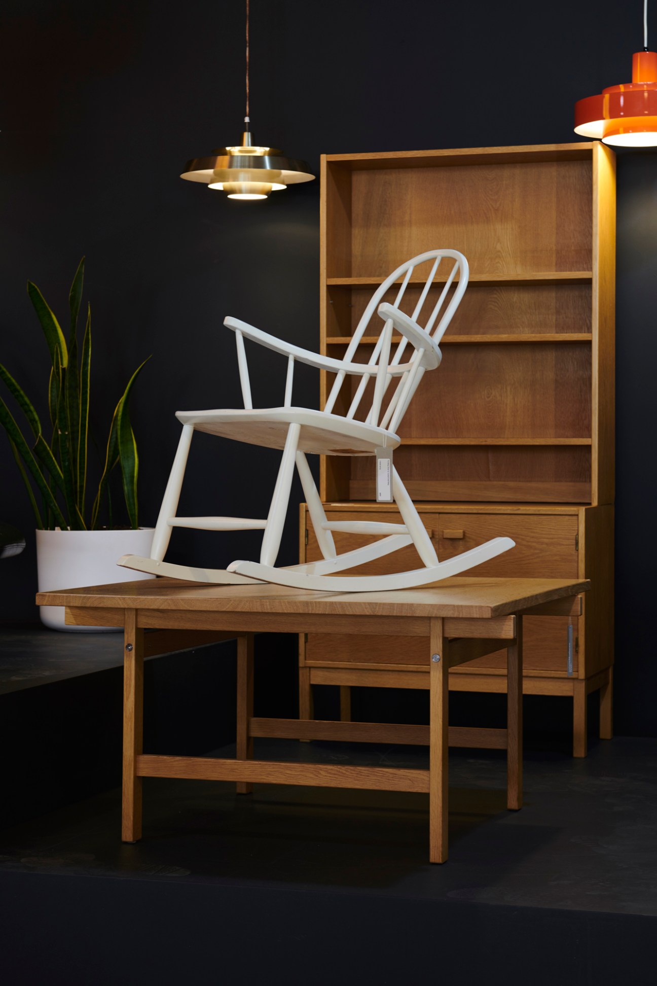Mid-century modern furniture display featuring a white rocking chair, wooden desk, and shelving unit under pendant lighting.