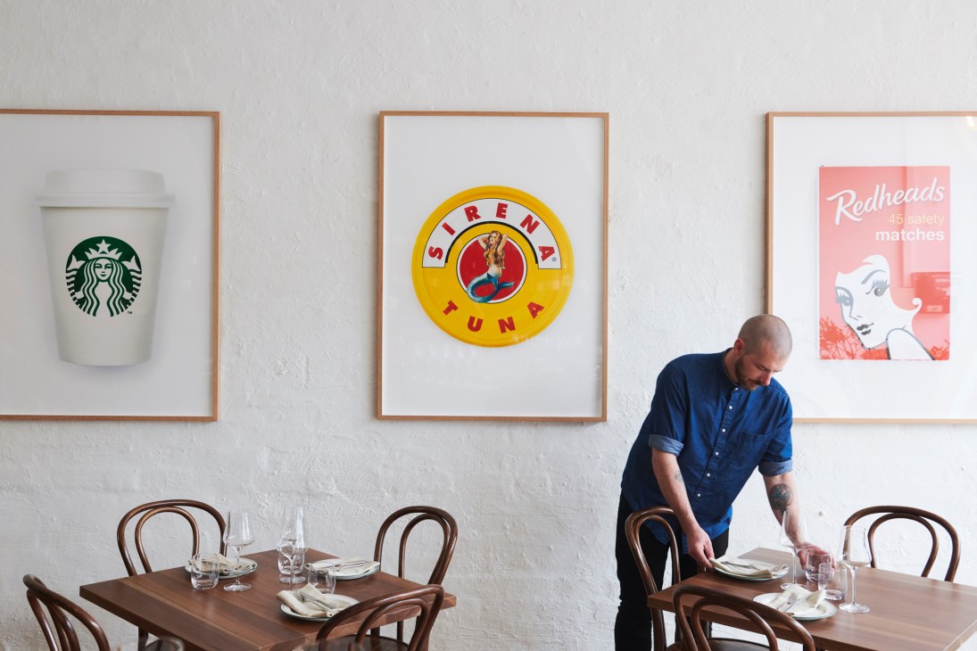 Man in blue shirt setting table at Vex restaurant with framed artwork on wall behind