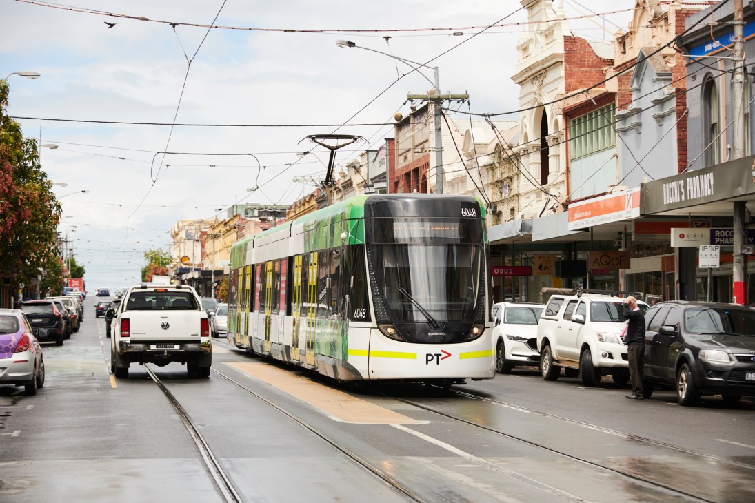 A tram travels along High Street in Northcote, Melbourne, passing Victorian buildings and parked cars.