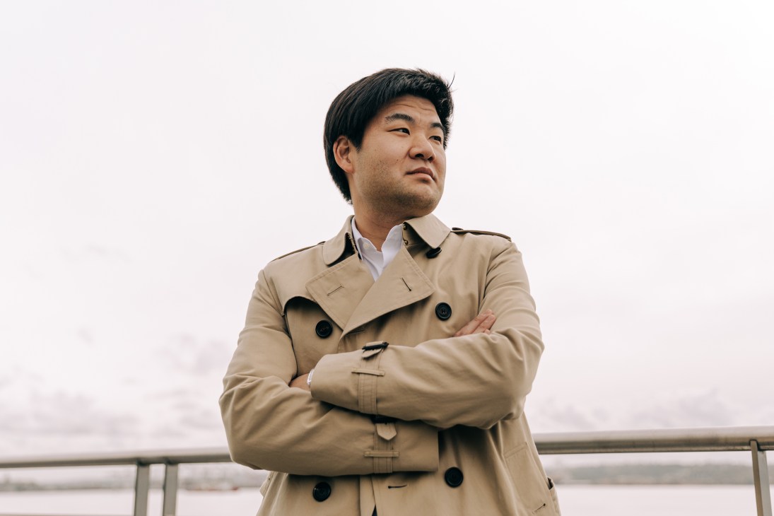 Man in beige trench coat with arms crossed standing outdoors against cloudy sky.