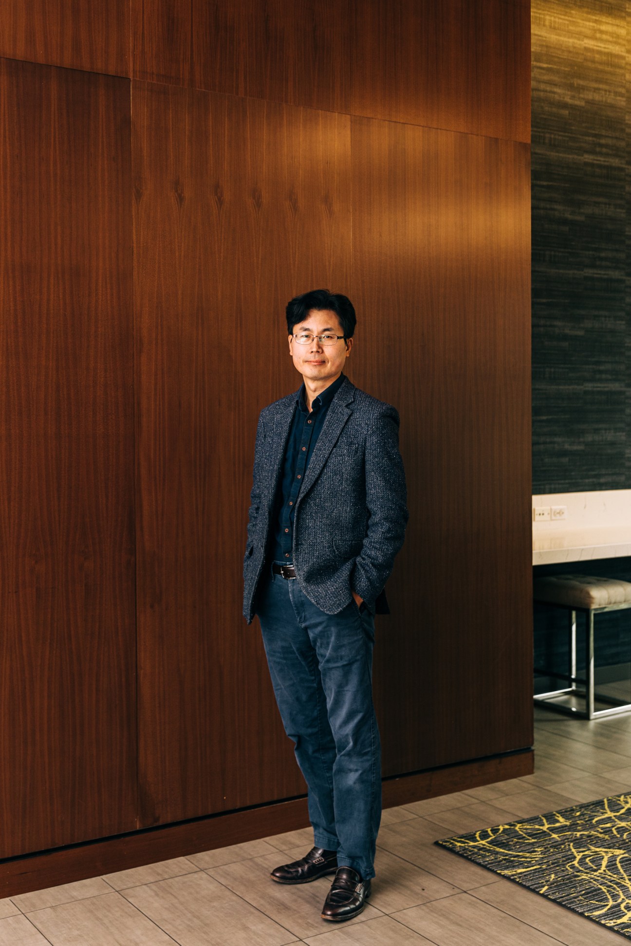 Man in glasses and grey blazer standing against wooden wall panels in modern interior space.