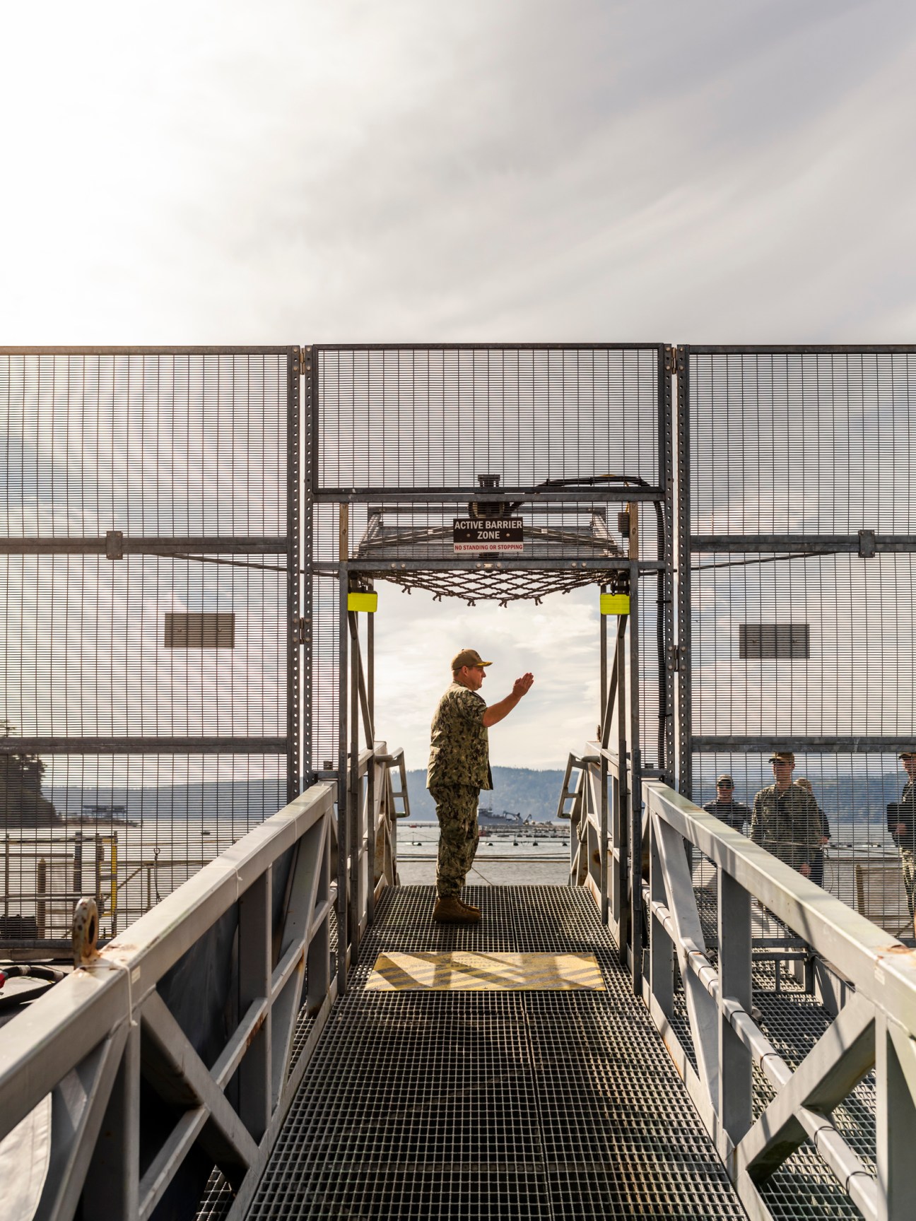 Military personnel in camouflage uniform standing on a metal walkway at a naval dock with water and mountains visible through a gate.