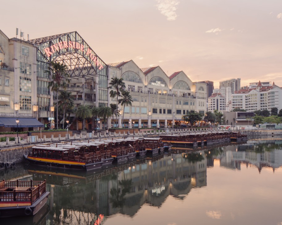 Projector X: Riverside waterfront complex in Singapore at dusk with boats moored below.