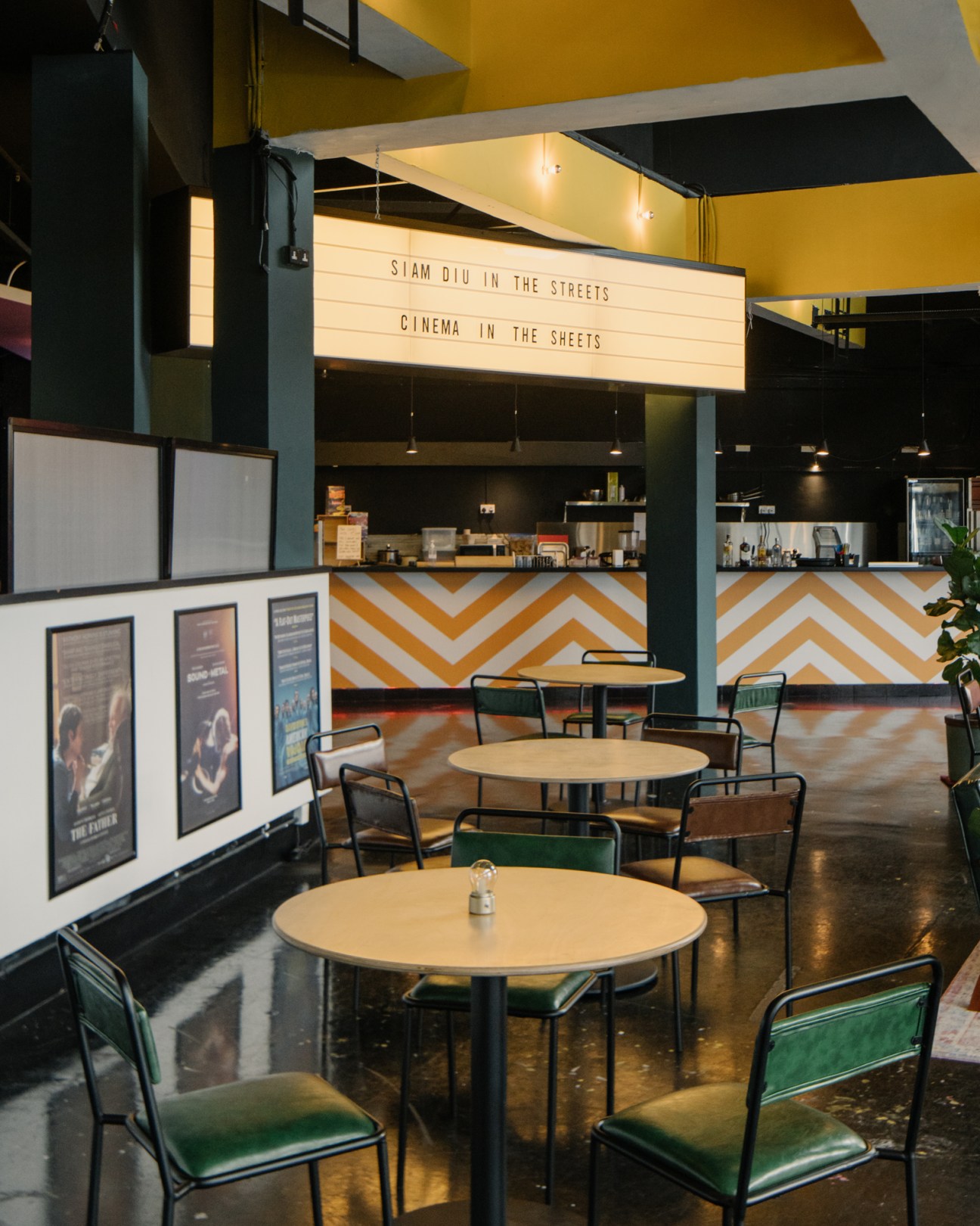 Interior of Projector X: Riverside cinema cafe with round tables, chairs, and yellow and white geometric patterned counter with illuminated marquee sign.