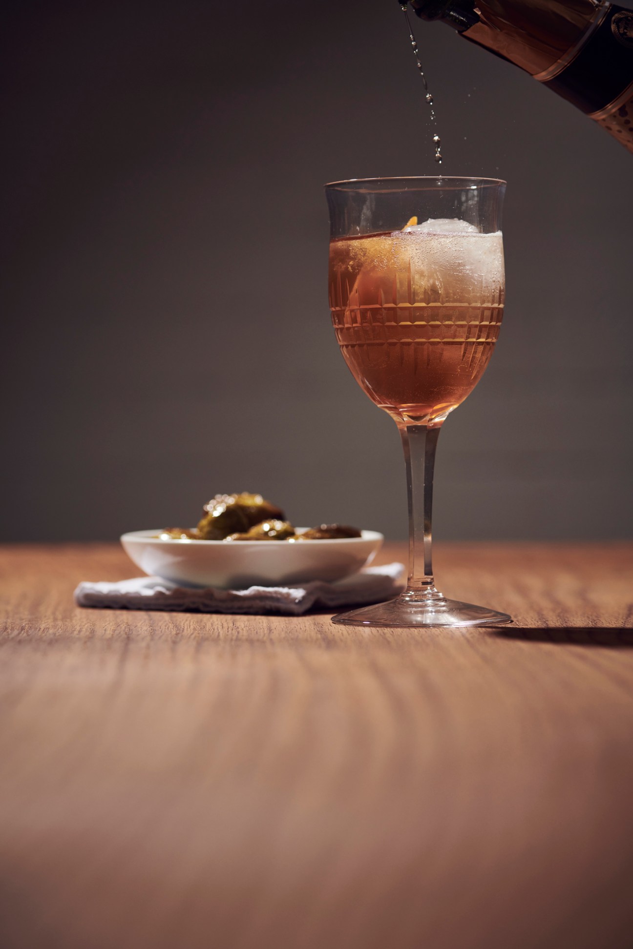 Golden cocktail in wine glass with large ice cube and orange garnish, bubbles rising from champagne