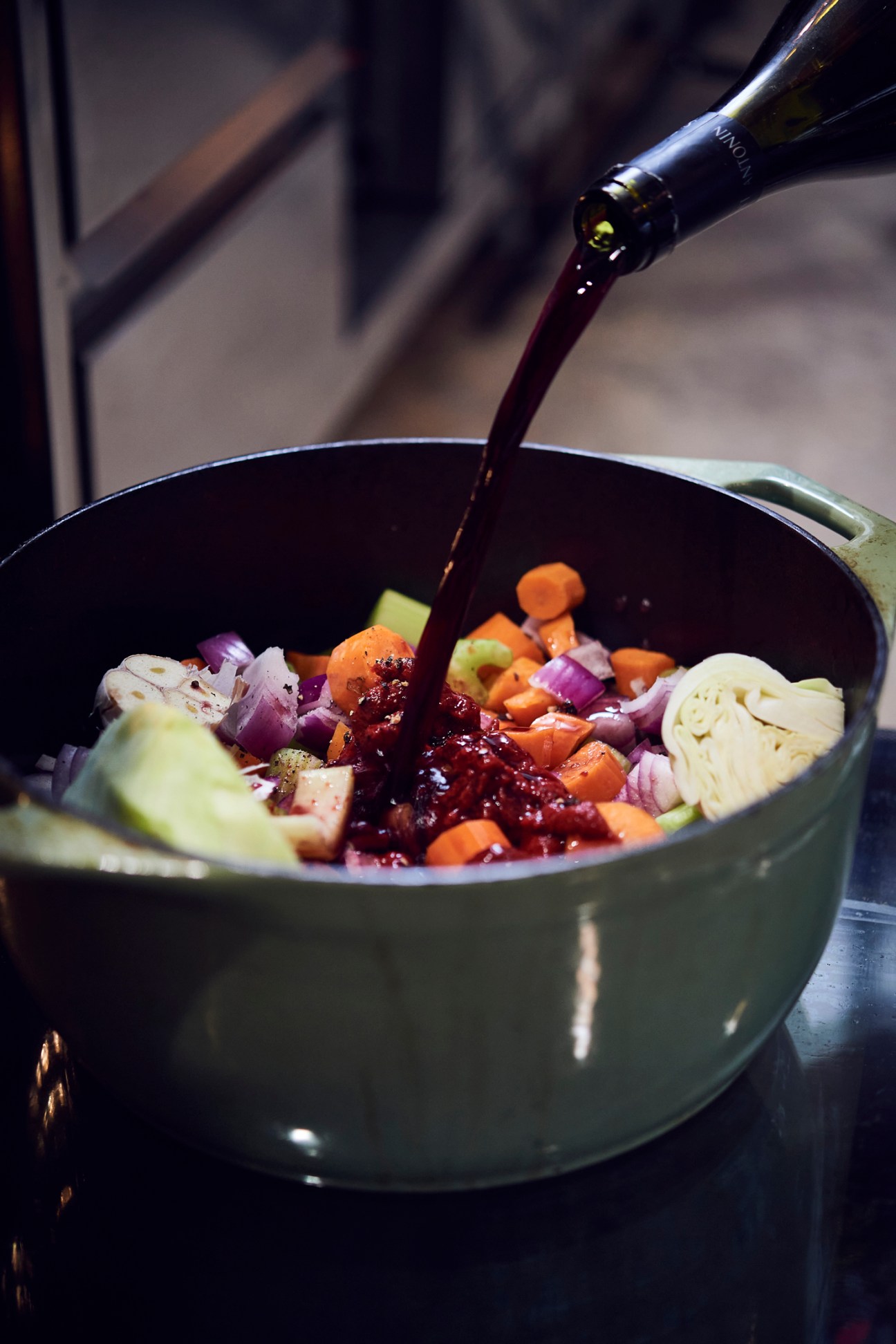 Red wine being poured from a bottle into a large pot containing chopped vegetables for cooking