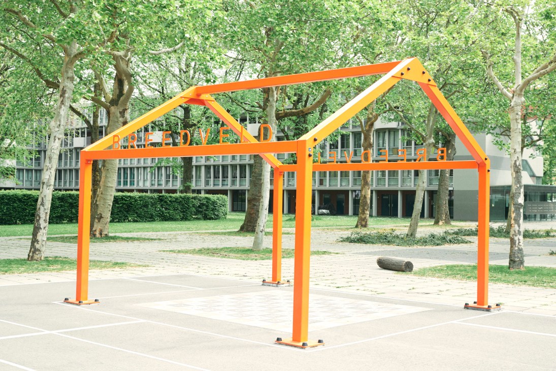Bright orange house-shaped metal structure in an urban plaza with trees and buildings in background.
