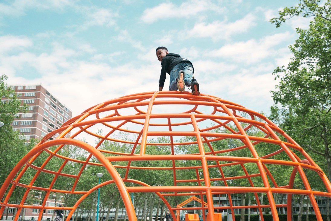 A child climbing on a bright orange steel-framed dome structure in an urban playground with residential buildings in the background.