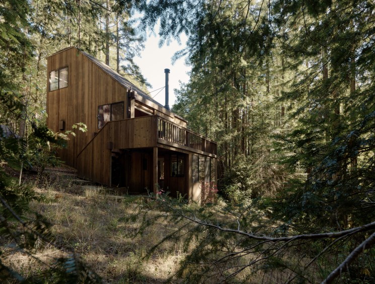 cabin on a wooded hillside seen through dense trees.