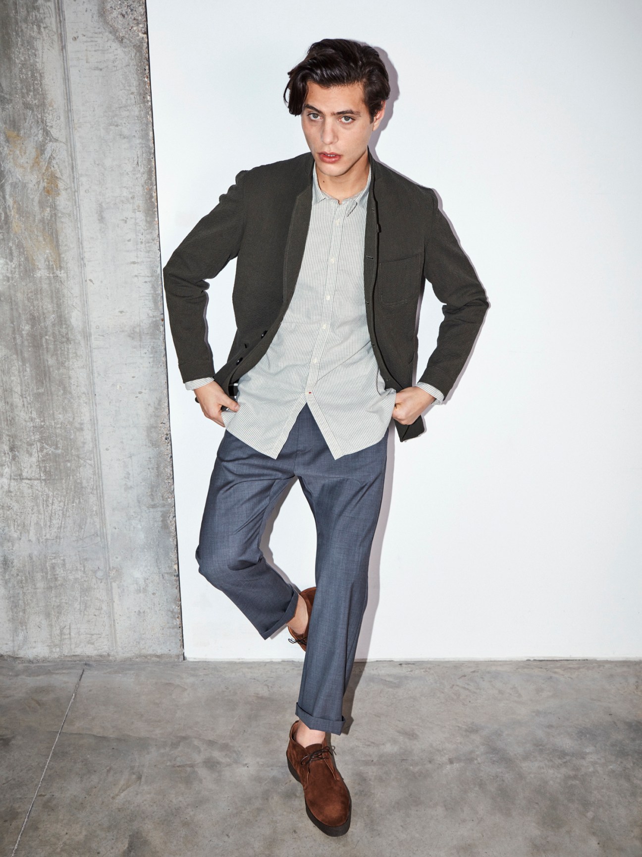 Man in dark blazer, striped shirt and blue trousers posing against concrete wall