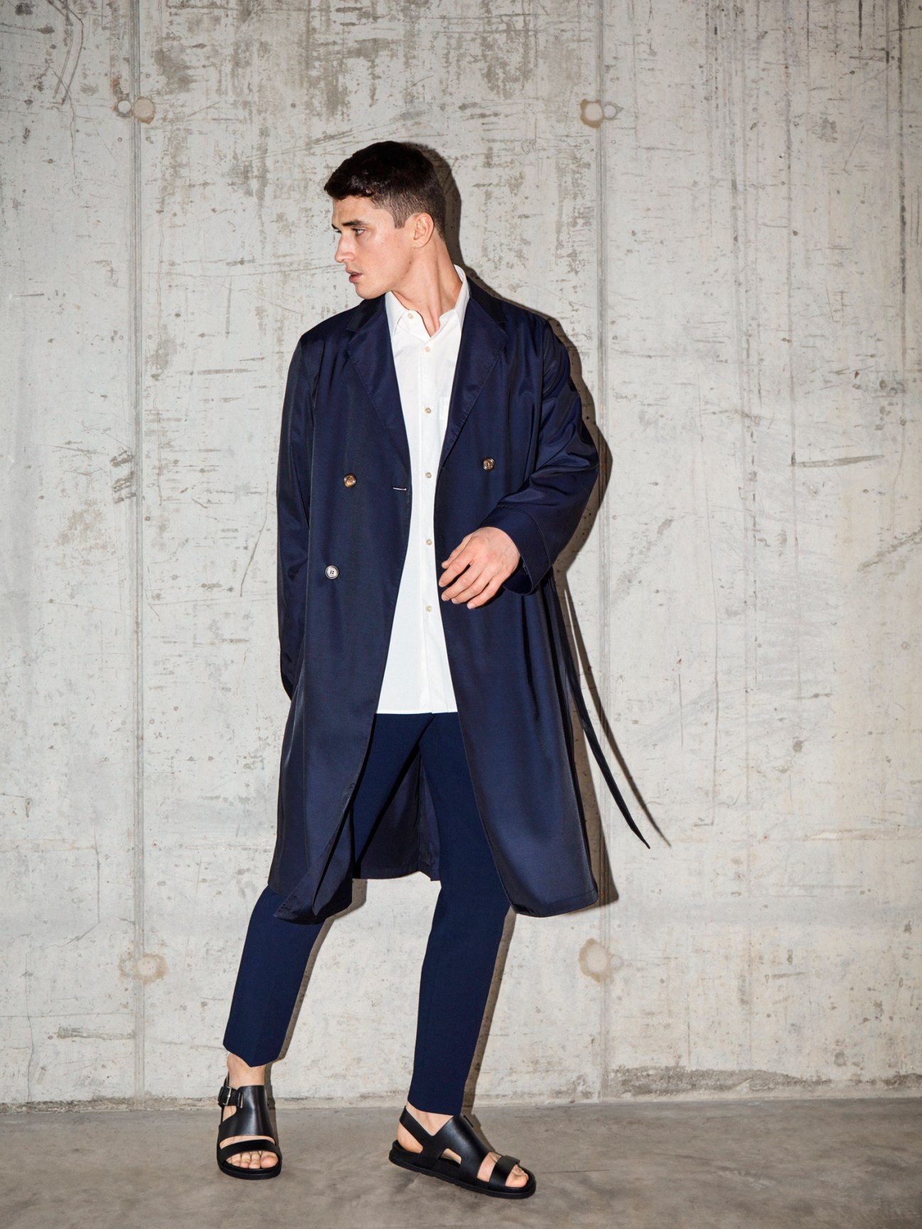 Man in navy Bally coat, Studio Nicholson shirt, Giorgio Armani trousers and Hermès sandals against concrete wall