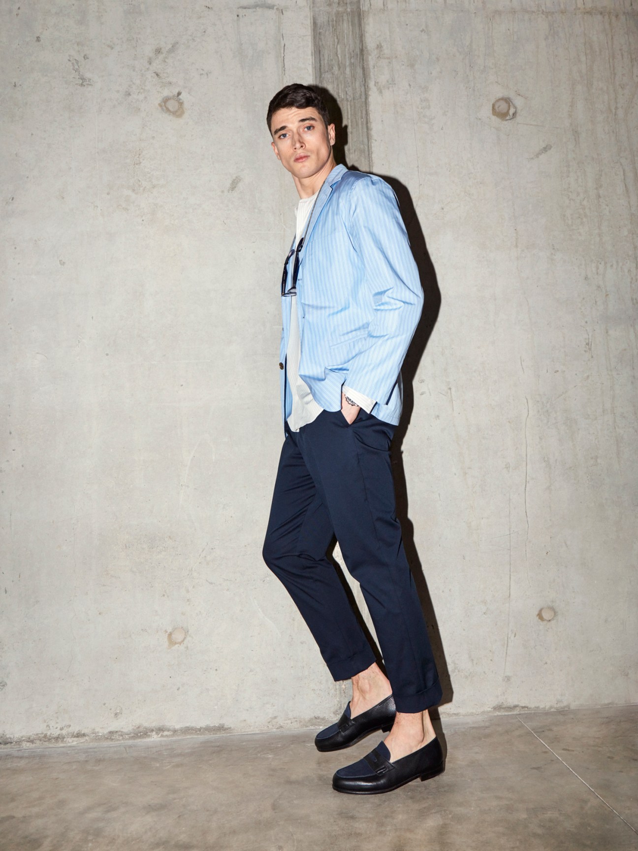 Man in light blue striped blazer, white t-shirt, navy trousers and black loafers leaning against concrete wall