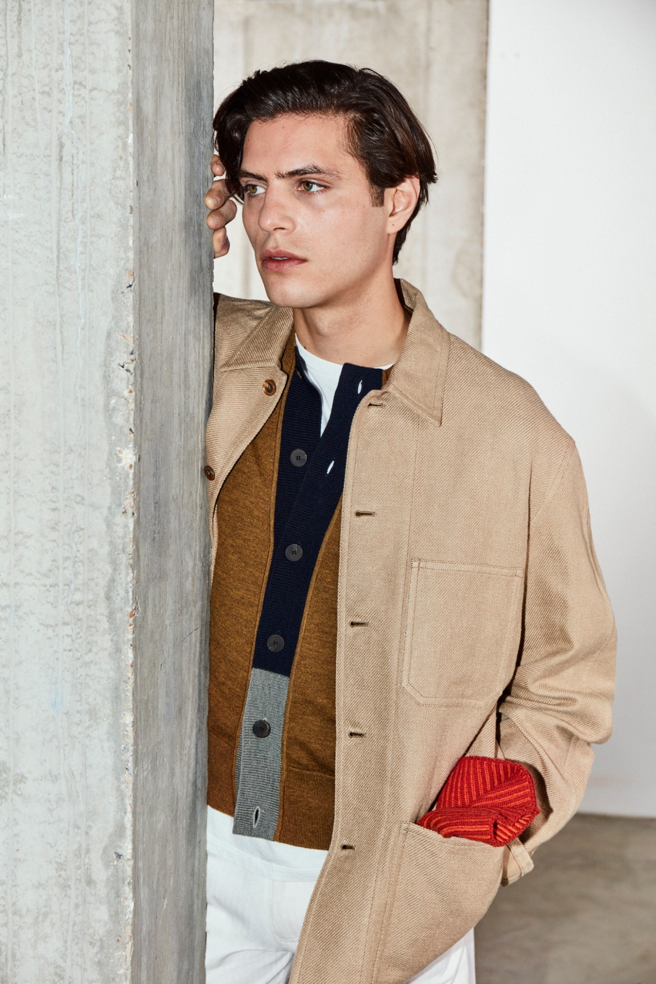 Man in beige jacket and layered clothing leaning against concrete wall