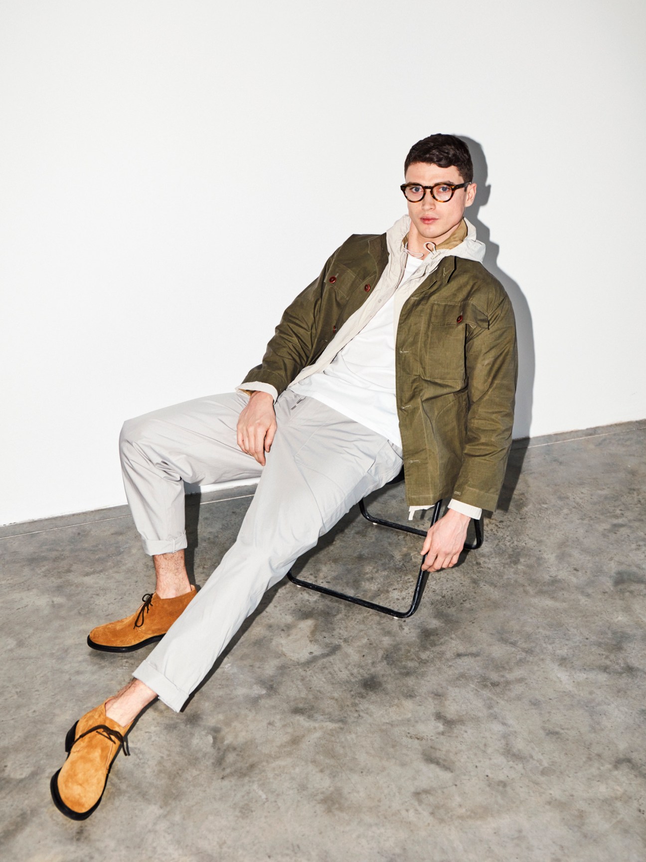 Man in olive jacket, white t-shirt, light trousers and tan suede shoes sitting on modern chair