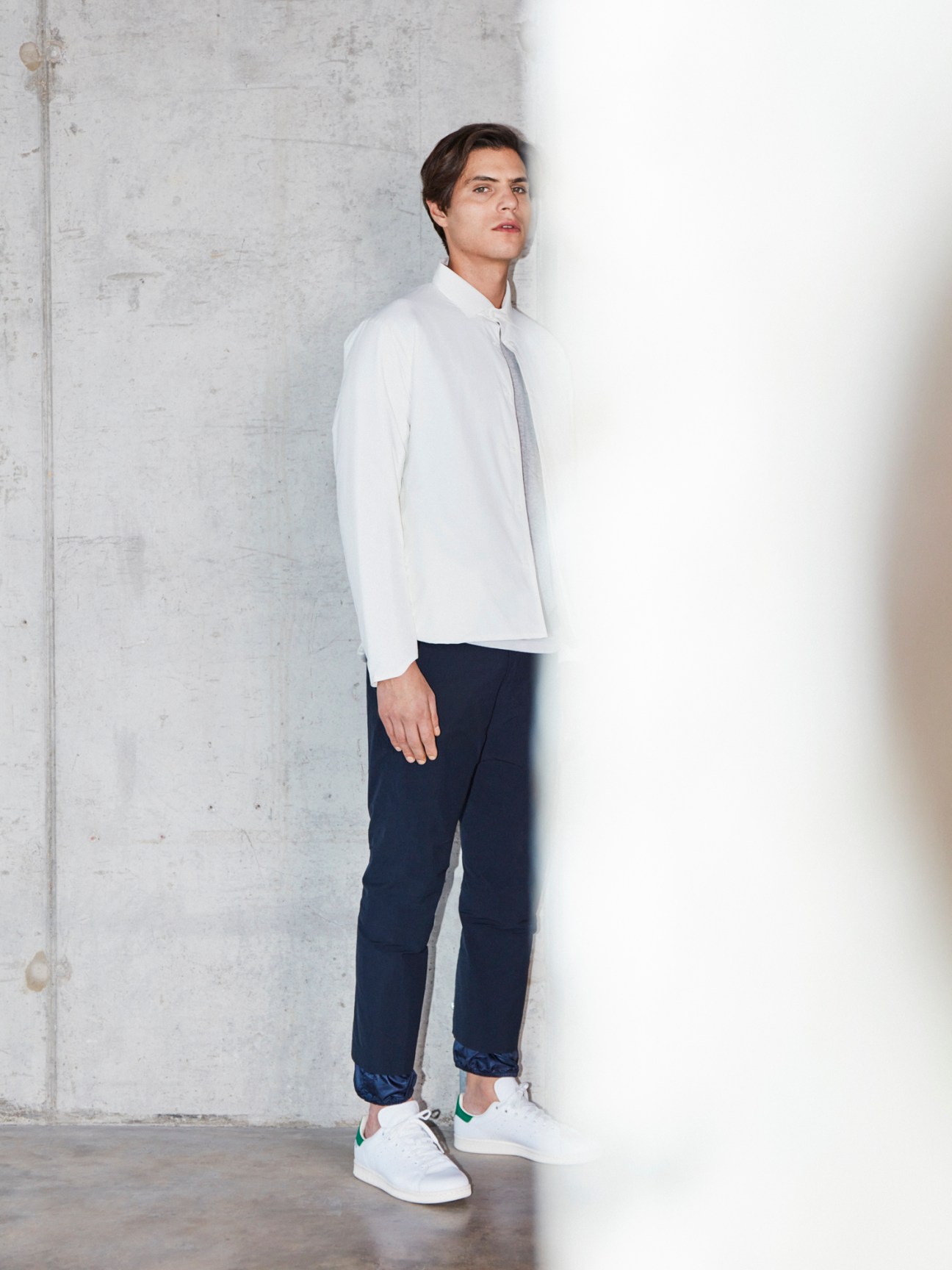 Man in white jacket and navy trousers standing against concrete wall wearing white sneakers