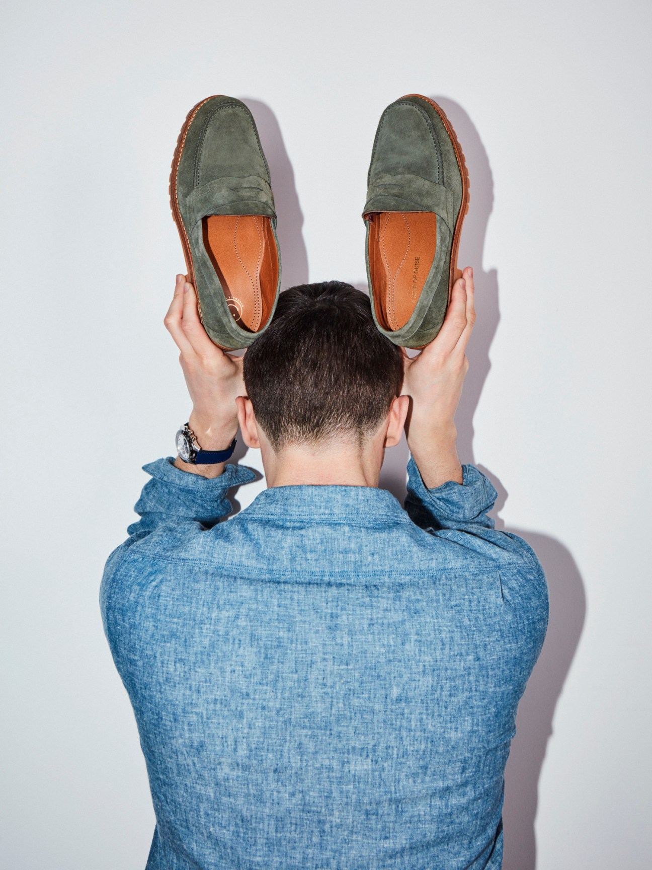 Person in blue chambray shirt holding olive green suede loafers up to their head like ears