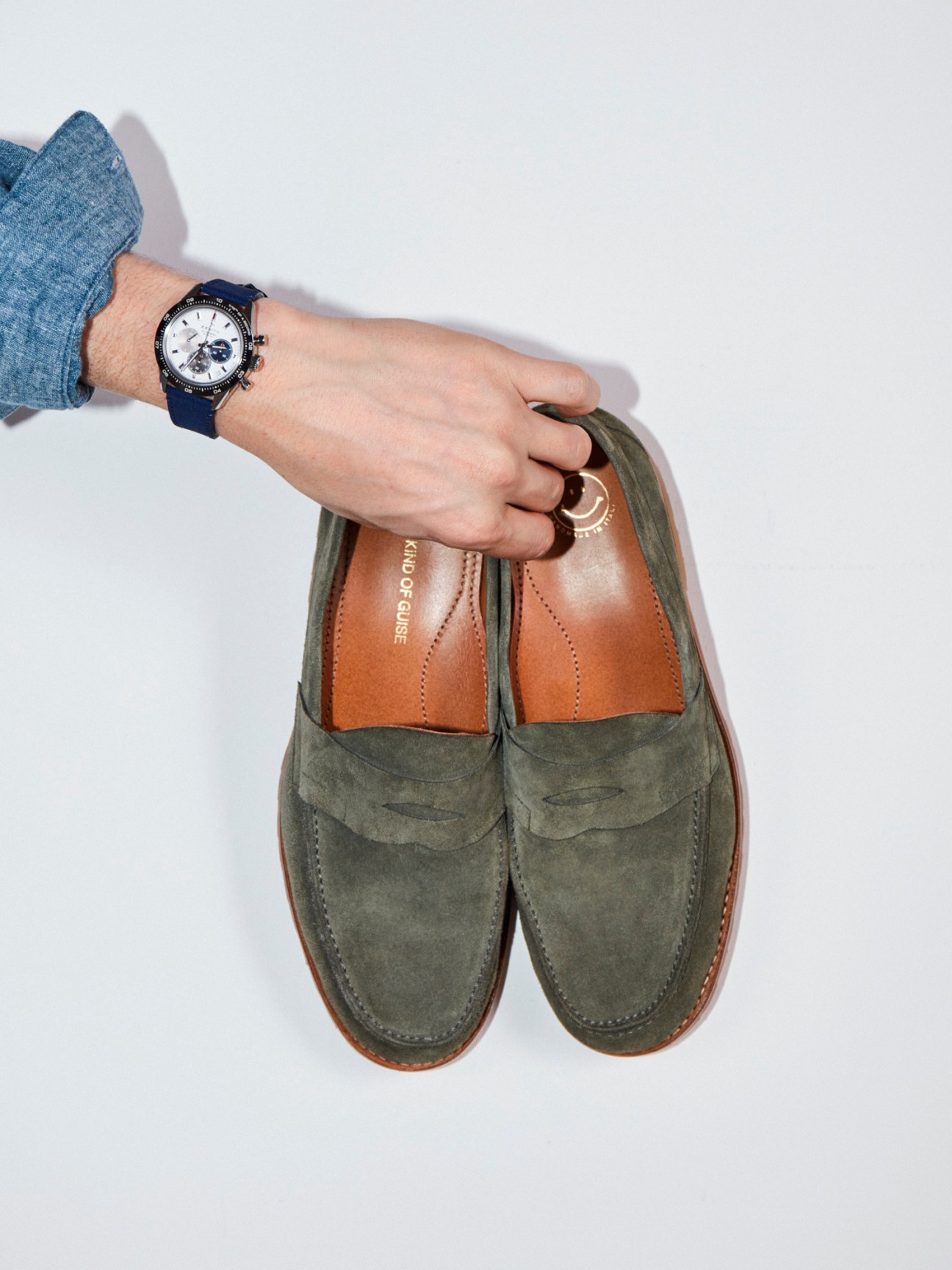 Hand wearing watch reaching for olive green suede penny loafers with brown leather lining