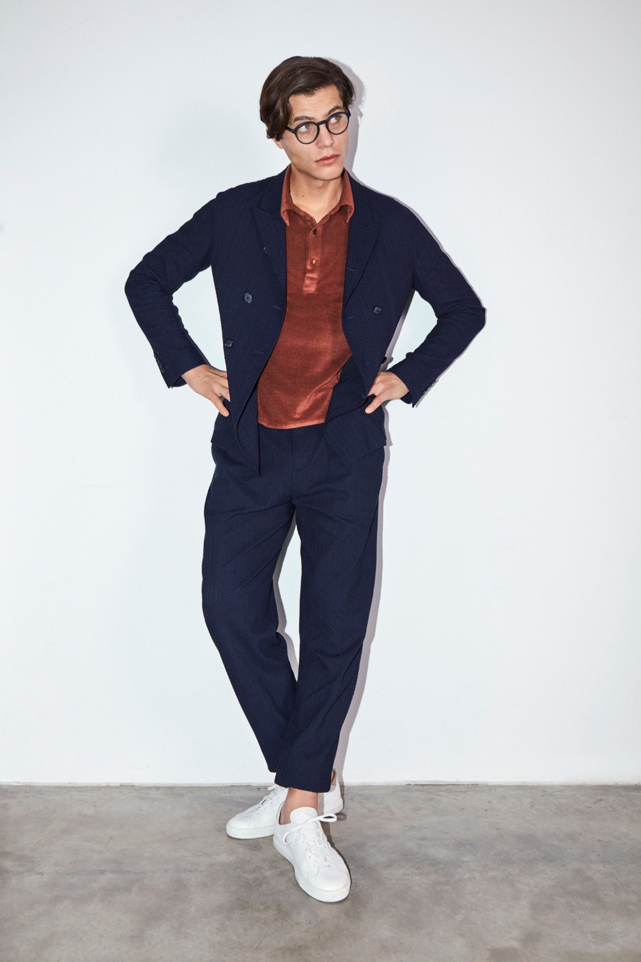 Person in navy Giorgio Armani suit with rust polo shirt, white trainers and glasses posing against white wall