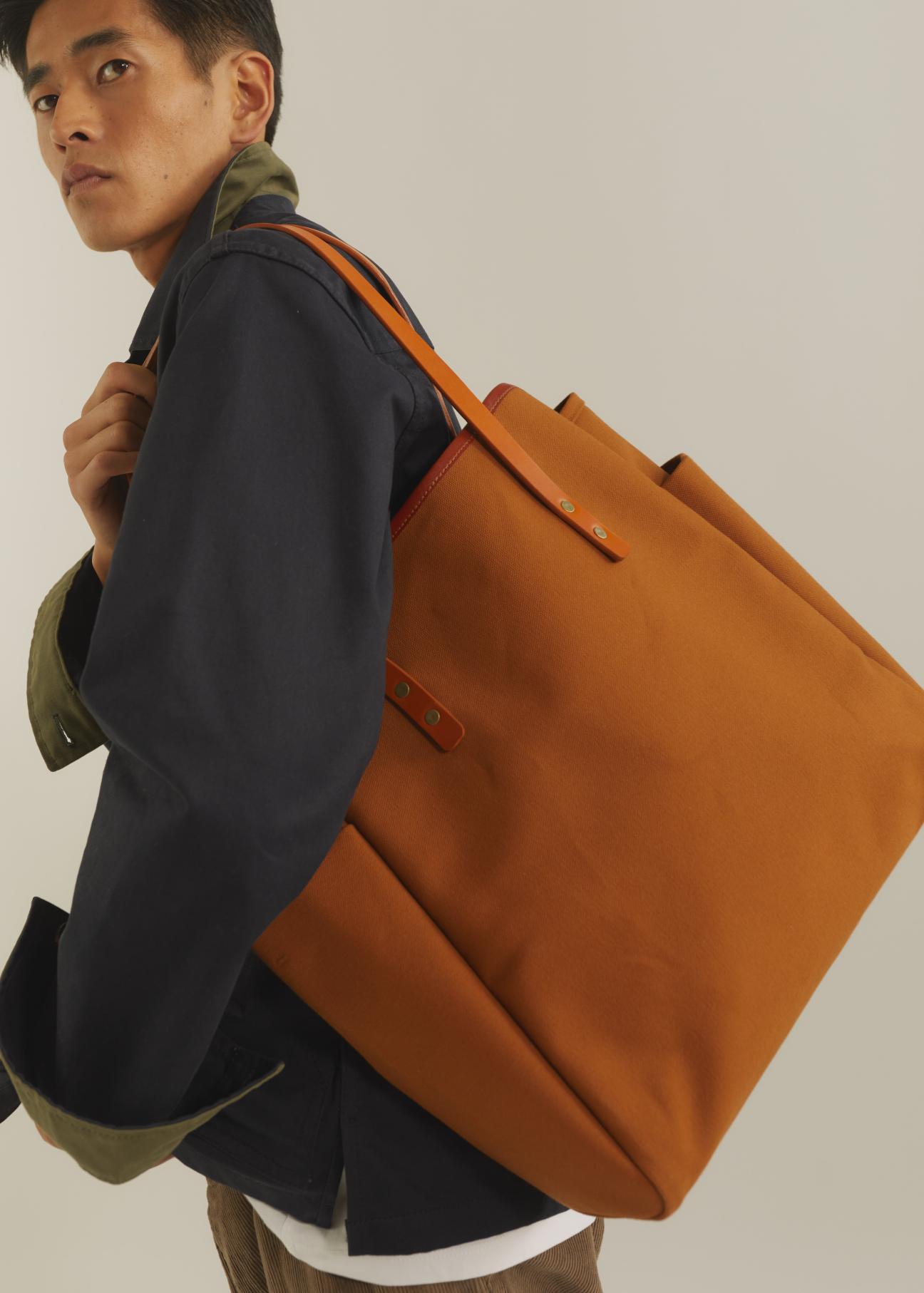 Orange waxed canvas tote bag with leather straps being carried by person in navy jacket