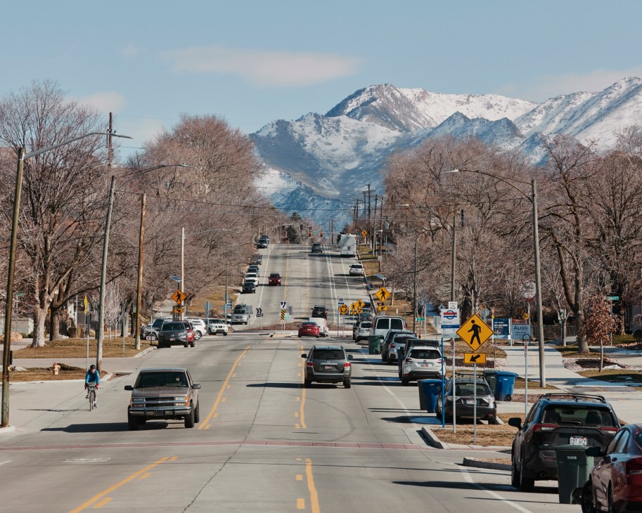 Street view looking uphill toward snow-capped mountains with cars, power lines, and bare winter trees