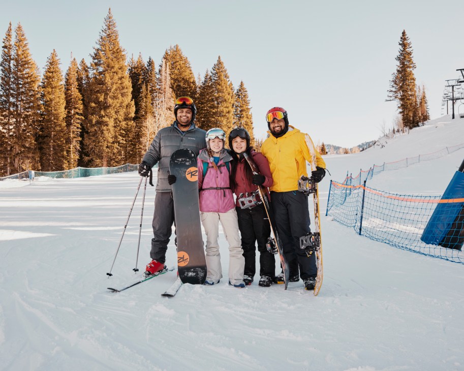Four friends posing together on snowy ski slopes with skis and snowboards, wearing winter gear and goggles