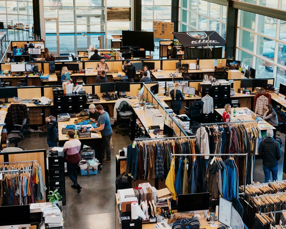 Open office workspace with people at desks, clothing racks displaying garments, and large windows