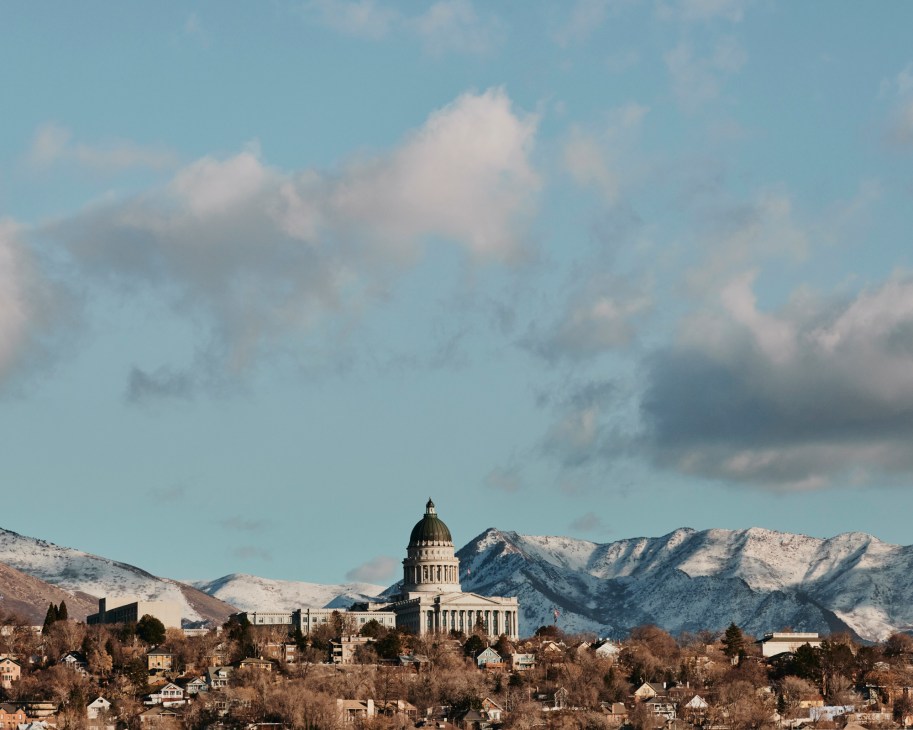 Utah State Capitol building with domed roof against snow-covered Wasatch Range mountains under cloudy sky