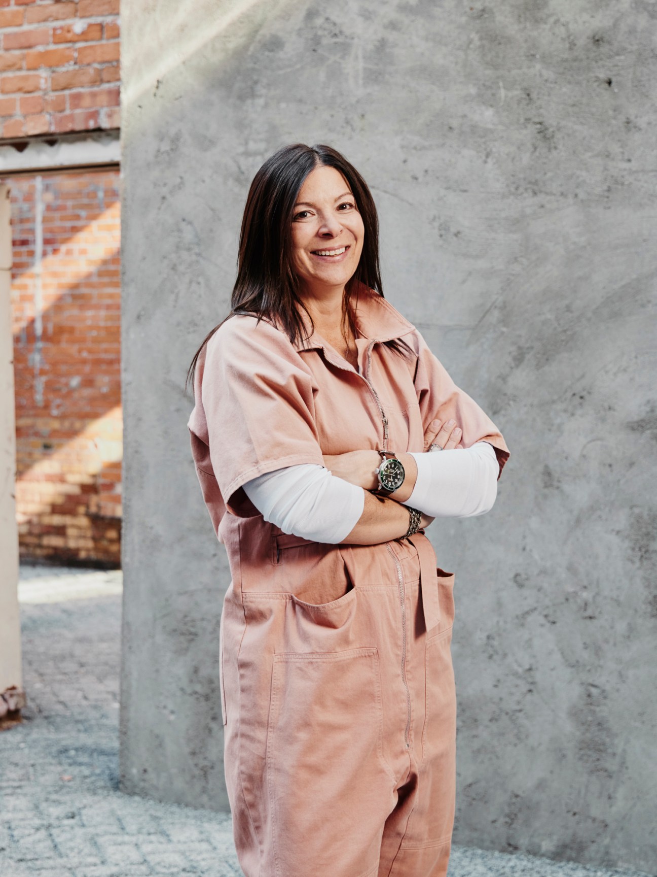 Ellen Winkler standing with arms crossed, wearing a pink coat and watch, smiling at camera