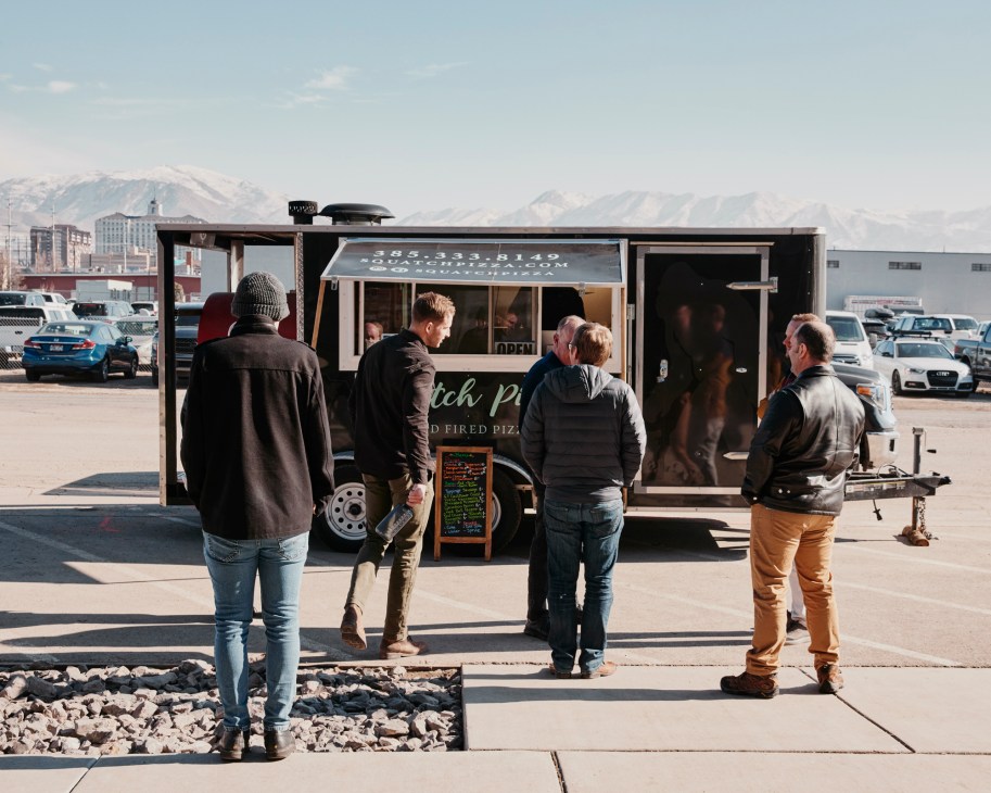 People standing in line at a food truck in a parking lot with snow-capped mountains in the background