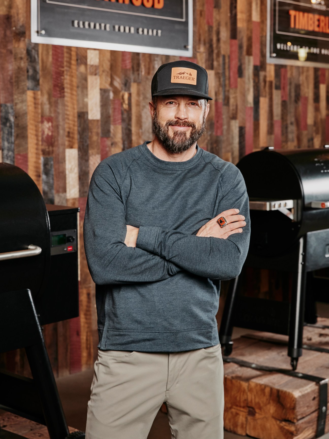 Jeremy Andrus of Traeger standing with arms crossed in front of grills and rustic wood wall