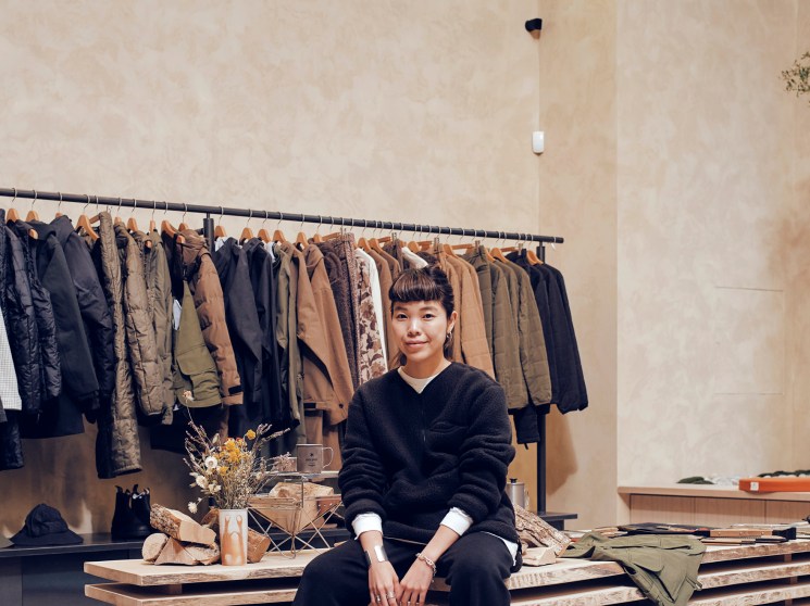 A young woman with a short fringe sits in front of a rail of earth-toned jackets in a clothing boutique.