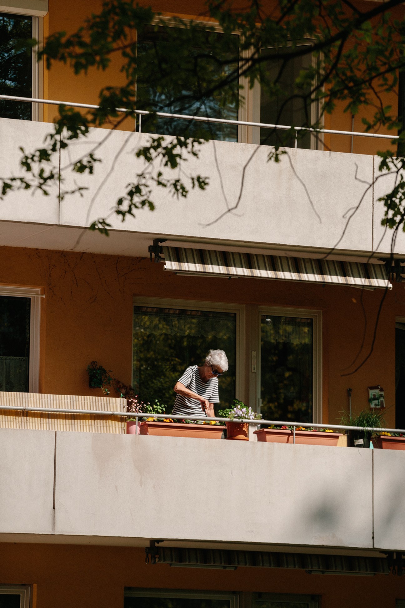 Elderly woman in striped shirt tends to potted flowers on a sunny apartment balcony.