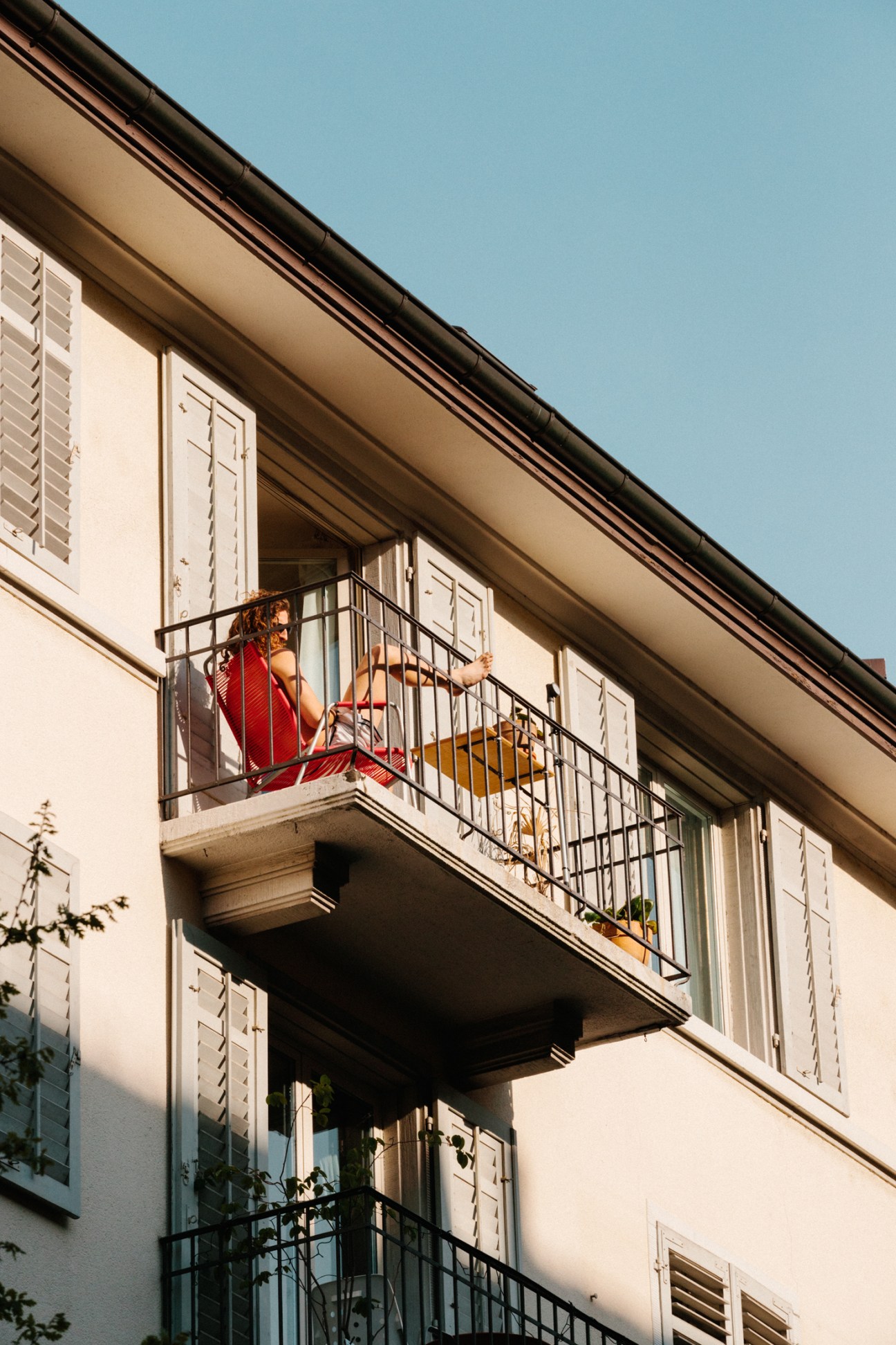 Woman in red dress standing on a balcony with a yellow chair against a cream-colored building facade.