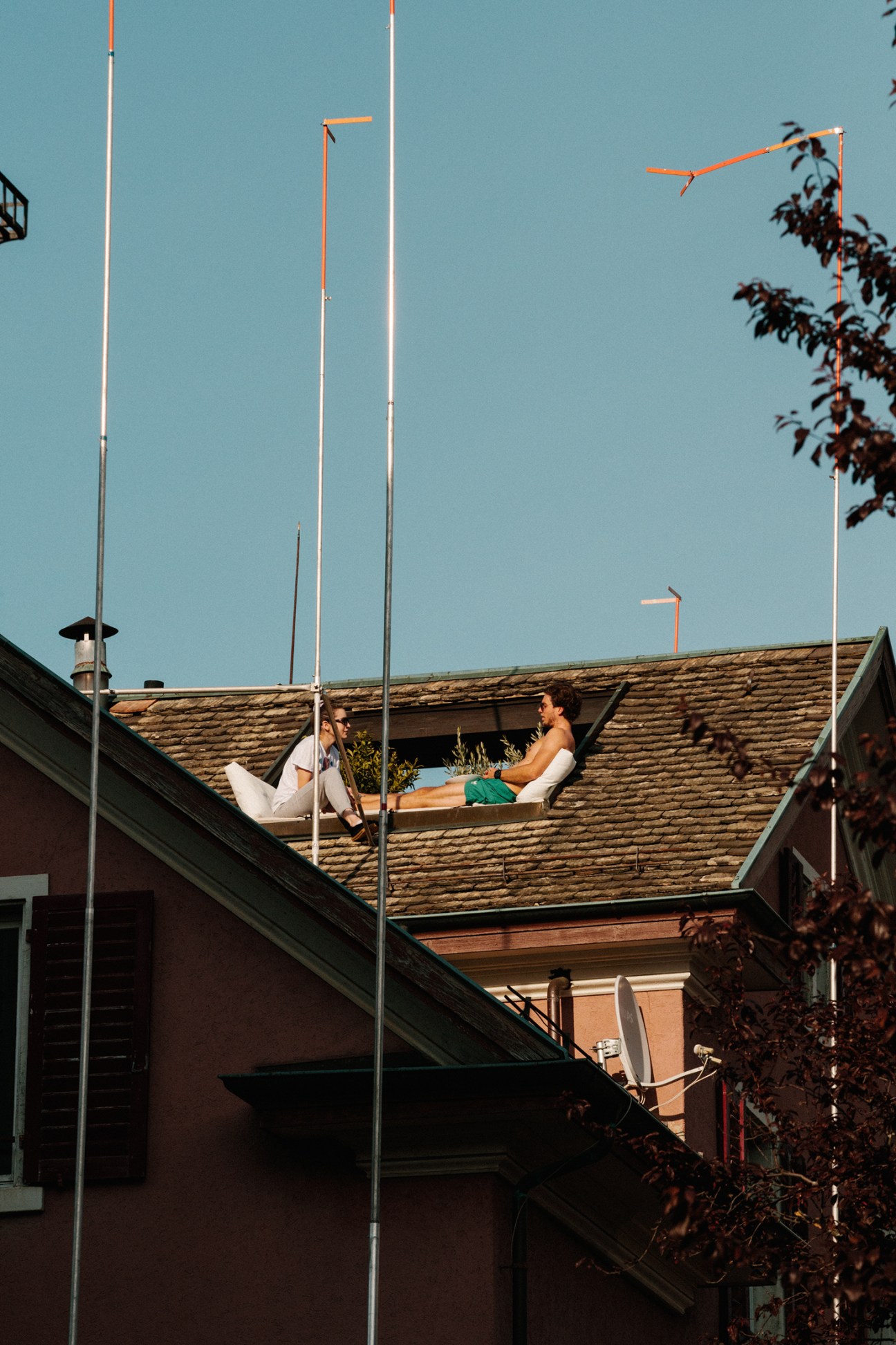 Two people sit on a rooftop with clear sky above, surrounded by metal poles and autumn foliage.
