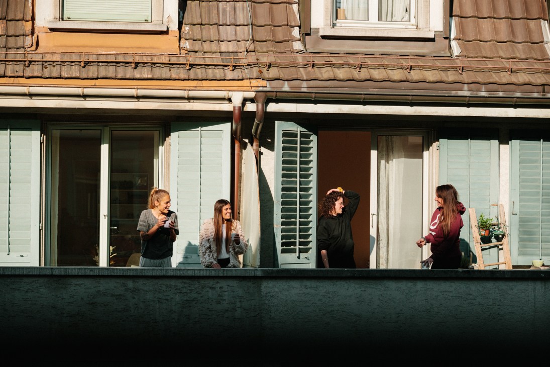 Five people stand at windows and on a balcony of a modern building with white shutters during daytime.